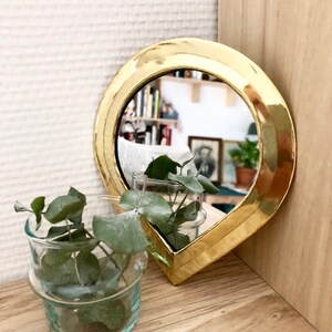 May include: A gold-colored teardrop-shaped mirror with a beveled edge. The mirror is reflecting a bookshelf and a plant.