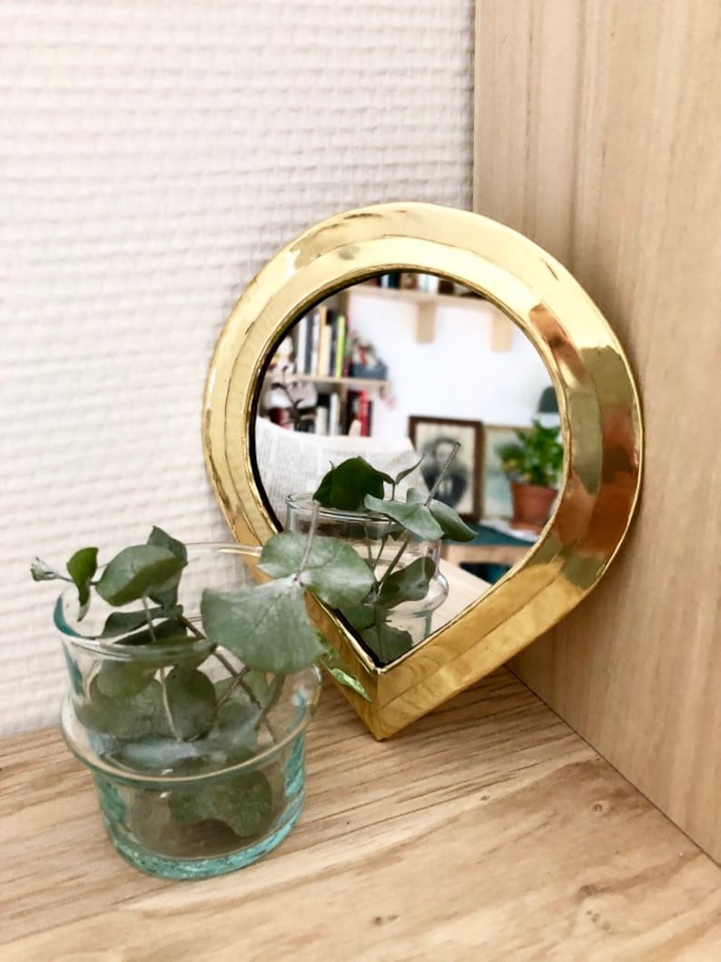 May include: A gold-colored teardrop-shaped mirror with a beveled edge. The mirror is reflecting a bookshelf and a plant.