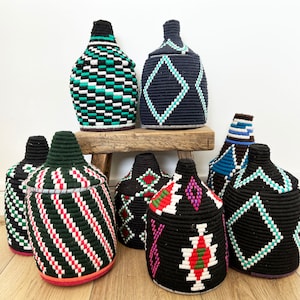 Puede incluir: Una colección de cestas de almacenamiento tejidas con tapas. Las cestas presentan varios patrones geométricos en colores como negro, blanco, verde, rojo y azul. Se exhiben sobre un taburete de madera contra una pared blanca.