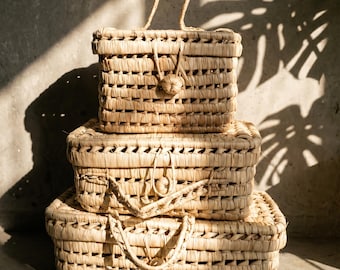 Puede incluir: Tres cestas rectangulares tejidas apiladas. Las cestas están hechas de un material natural marrón claro y tienen asas. La cesta superior tiene un asa, las dos inferiores tienen dos. Las cestas están en un espacio bien iluminado.