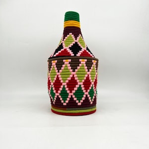 Può includere: Un contenitore decorativo a forma di bottiglia con un design intrecciato. Il contenitore presenta un motivo di diamanti rossi, verdi, rosa e neri. Il collo è verde e giallo e la base è rossa e marrone. Il contenitore è probabilmente fatto a mano.