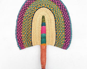 May include: Handwoven fan with a rainbow design. The fan features a natural straw center, surrounded by bands of pink, teal, orange, and purple. The handle is wrapped in brown leather with a dark blue loop at the end. A decorative and functional item.