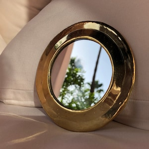 Puede incluir: Un peque&ntilde;o espejo redondo de color dorado con un borde biselado. El espejo refleja una imagen borrosa de &aacute;rboles y un cielo azul.