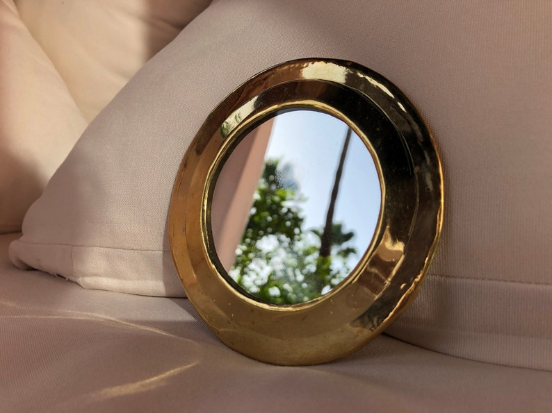 Puede incluir: Un peque&ntilde;o espejo redondo de color dorado con un borde biselado. El espejo refleja una imagen borrosa de &aacute;rboles y un cielo azul.