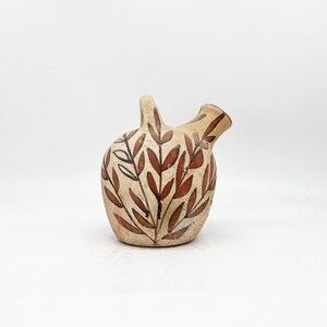 May include: A beige ceramic pitcher with a handle and spout. The pitcher is decorated with a pattern of brown and red leaves. The design is painted on the surface of the pitcher. The pitcher is on a white background.