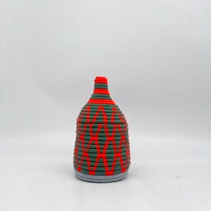 May include: A woven basket with a bottle-like shape. The basket is primarily green with a red zig-zag pattern and a red top. The base is a light gray color. The basket is made of a woven material.