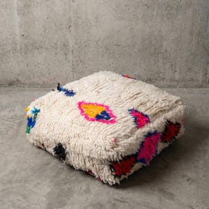 May include: A square, off-white pouf with a shaggy texture. It features colorful diamond-shaped patterns in pink, yellow, blue, and red. The pouf is placed on a concrete surface, showcasing its decorative design.