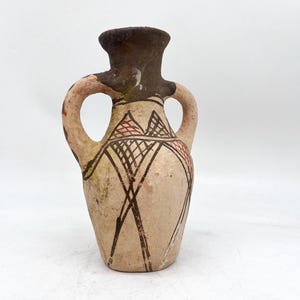 May include: A small, light-colored ceramic vessel with two handles and a dark brown neck. The body features hand-painted geometric patterns in black and reddish-brown, consisting of intersecting lines and cross-hatched triangular shapes.