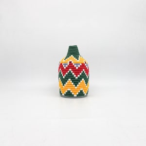 Puede incluir: Una pequeña cesta tejida de color verde, amarillo y rojo con un patrón geométrico. La cesta está hecha de fibras naturales y tiene una forma redonda.