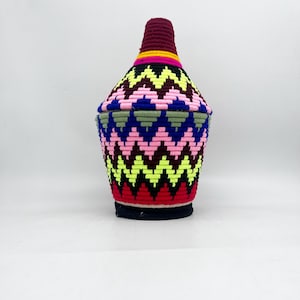 May include: A woven storage basket with a geometric pattern in shades of pink, red, yellow, blue, green, and black. The basket has a round base and a narrow opening.