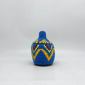 May include: A blue woven basket with a lid. The basket is decorated with yellow, red, and green geometric patterns. The lid has a rounded top. The basket is sitting on a white surface.