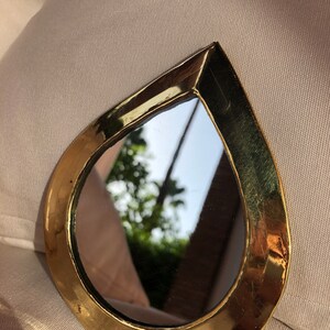 May include: A small, teardrop-shaped mirror with a gold-colored frame. The mirror reflects a blurry image of trees and a sky.