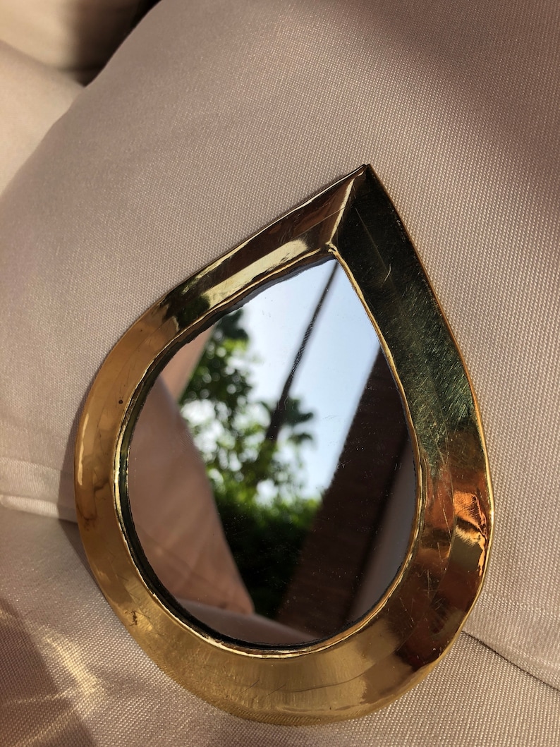 May include: A small, teardrop-shaped mirror with a gold-colored frame. The mirror reflects a blurry image of trees and a sky.