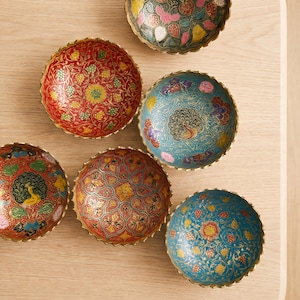 May include: Six decorative, round bowls with scalloped edges, featuring intricate floral and paisley patterns in red, blue, and gold. The bowls are arranged on a light-coloured wooden surface. Each bowl has a unique design.