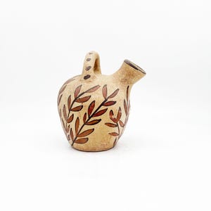 May include: A small, beige ceramic pitcher with a handle and spout. The pitcher is decorated with brown leaf patterns. The handle has small brown dots. The spout is dark brown.