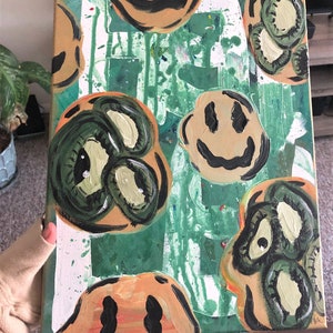 May include: A green and orange canvas painting featuring a variety of smiley faces. Some of the faces are green with large eyes, while others are orange with black eyes and a black smile.