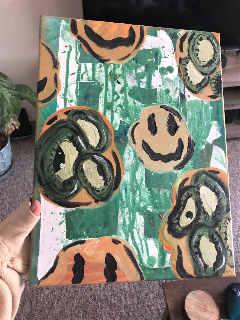 May include: A green and orange canvas painting featuring a variety of smiley faces. Some of the faces are green with large eyes, while others are orange with black eyes and a black smile.