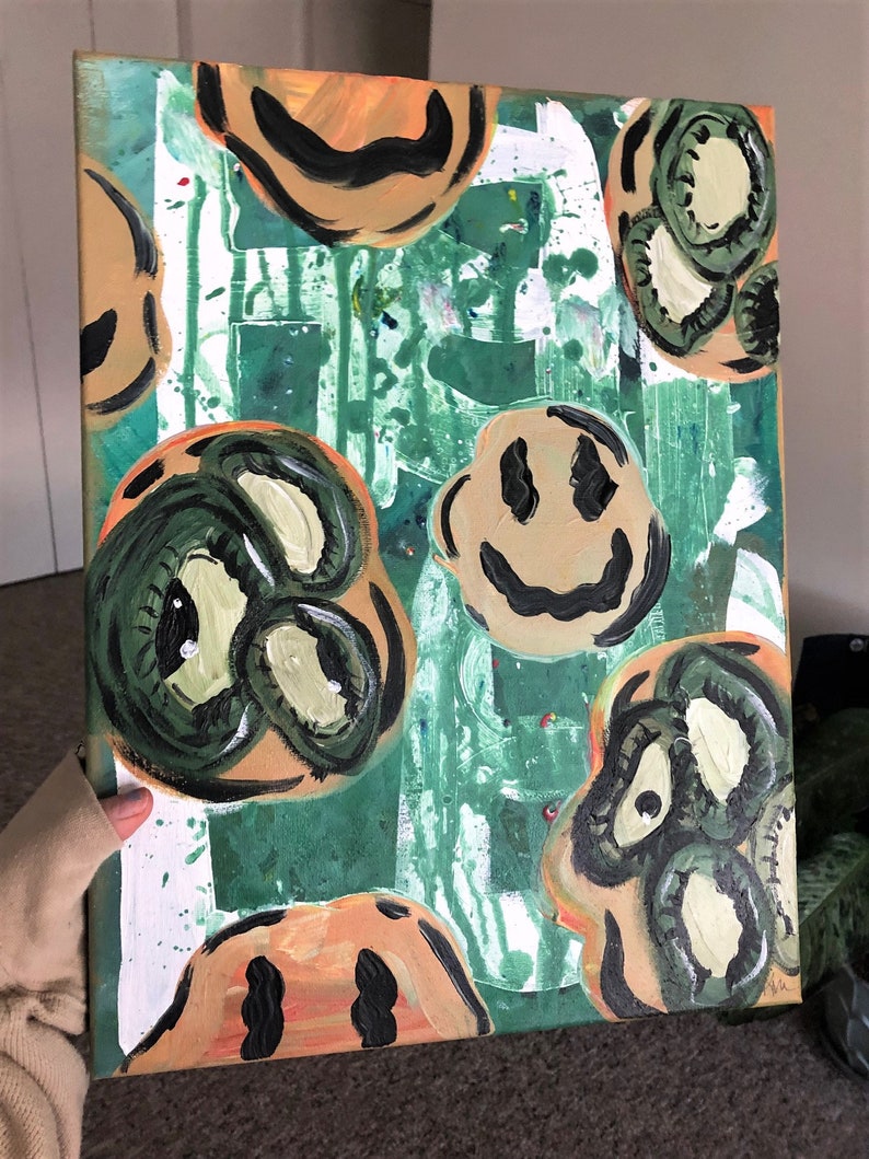 May include: Abstract painting on canvas with a green background and multiple smiley faces in various shades of green, yellow, and orange. The smiley faces are painted in a simple style with black outlines.