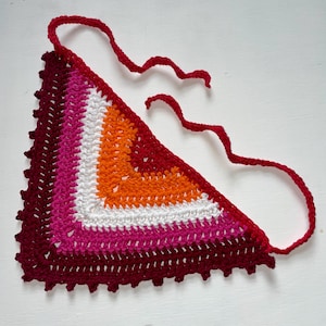 May include: A crocheted triangle in the colours of the lesbian pride flag. The design features stripes of dark red, pink, white, orange, and red, with a scalloped edge and red ties.