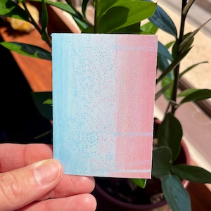 Puede incluir: Una pequeña tarjeta rectangular con un degradado de colores azul y rosa. La tarjeta está cubierta de pequeños puntos blancos.