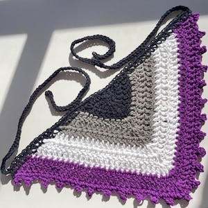 May include: A crocheted triangular scarf in the colours of the asexual pride flag: black, grey, white, and purple. The scarf has a black border and ties, and a scalloped purple edge.
