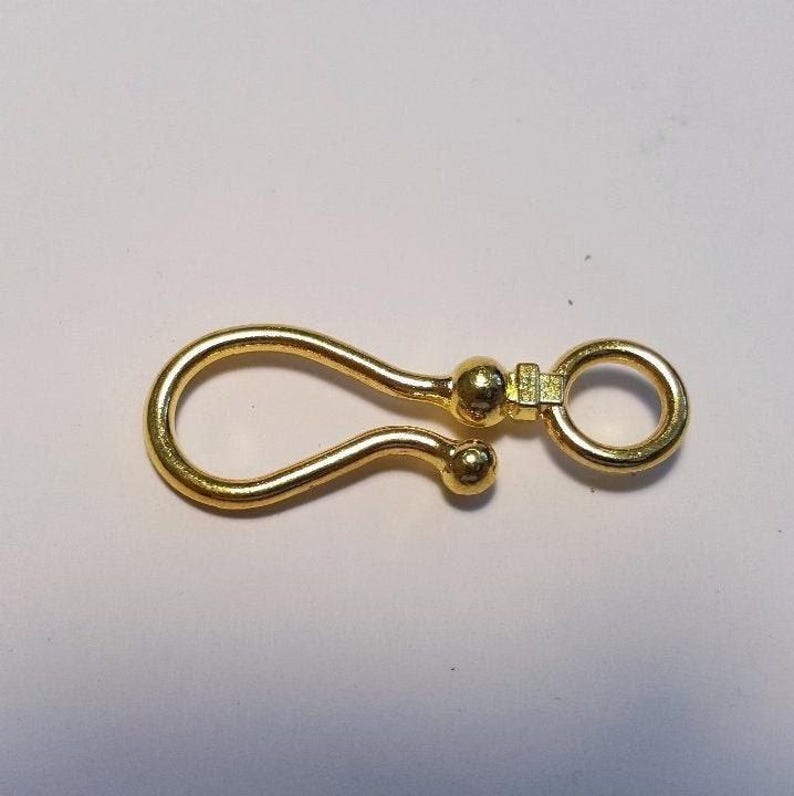 May include: A gold-toned metal hook clasp featuring a smooth, curved design with a rounded ball tip at the end of the hook. The clasp includes a small, circular ring attached to a central ball-shaped connector, providing a secure closure mechanism.
