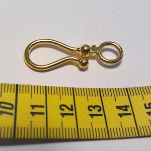 May include: A gold-colored metal hook clasp with a rounded loop and a swivel ring attachment, positioned above a yellow measuring tape that shows the object length is approximately 1.5 inches from end to end.