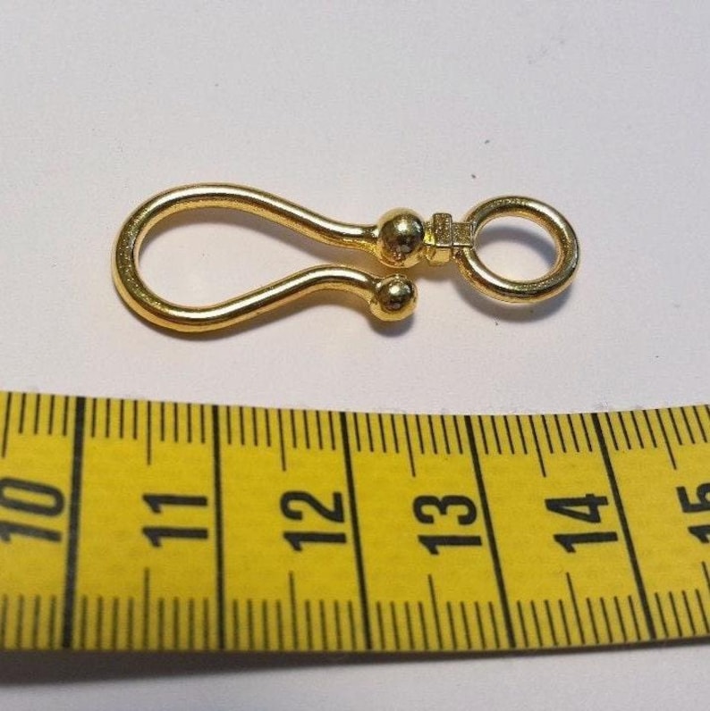 May include: A gold-colored metal hook clasp with a rounded loop and a swivel ring attachment, positioned above a yellow measuring tape that shows the object length is approximately 1.5 inches from end to end.
