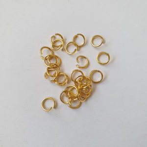 Puede incluir: Un grupo de anillos de salto dorados, algunos están apilados uno encima del otro, y otros están esparcidos sobre una superficie blanca.