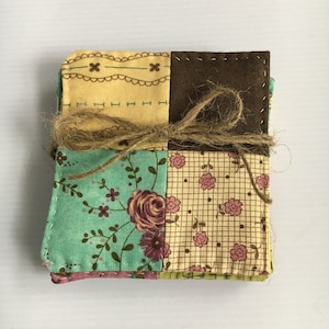 May include: A stack of square fabric coasters tied with twine. The coasters feature a patchwork design with floral and geometric patterns in shades of brown, yellow, green, and pink. The edges are stitched with white thread.
