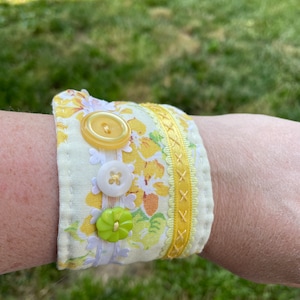 May include: A yellow and white fabric bracelet with floral print and yellow buttons. The bracelet is decorated with yellow and white trim.