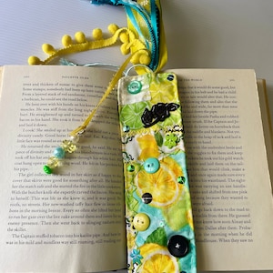 May include: A yellow and green fabric bookmark with a lemon print and a black rose. The bookmark is decorated with sequins and buttons. The bookmark is attached to a book with the text "OF THE WORLD" visible.