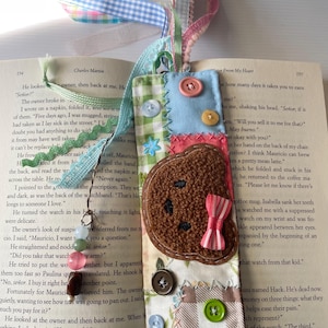 May include: A handmade fabric bookmark with a brown bear design, featuring a pink bow, colorful buttons, and a variety of patterned fabrics. The bookmark is attached to a book with the text "Letter From My Heart" visible.