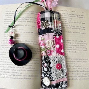 May include: A pink and black fabric bookmark with a black and white checkered pattern, a pink and white floral pattern, and a black and white paisley pattern. The bookmark has a small black hat with a pink band, a white button, and a silver star.