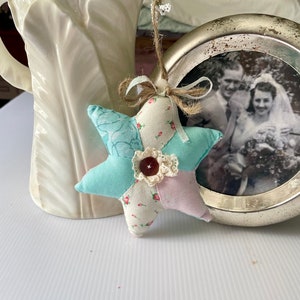 May include: A handmade patchwork star ornament in shades of blue, pink, and white with a floral print and a brown button in the center. The ornament is hanging from a twine string with a brown bow.