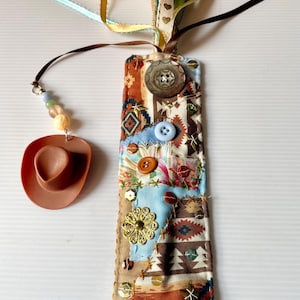 May include: A handmade bookmark with a patchwork design featuring various fabrics, buttons, and embellishments. It includes a small, brown cowboy hat charm and colorful ribbons. The bookmark is approximately 7 inches long.