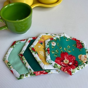 May include: Four fabric coasters in a hexagonal shape. The coasters are layered on top of each other. The top coaster is green with red flowers and white trim. The next coaster is yellow with a floral pattern and white trim. The next coaster is green with white trim. The bottom coaster is light green with white trim.