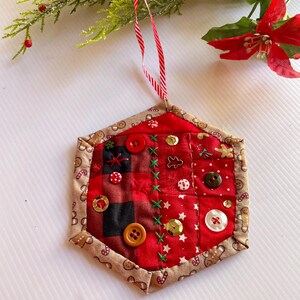 May include: A red and brown patchwork hexagon Christmas ornament with a gingerbread man border. The ornament is decorated with buttons, sequins, and embroidery.