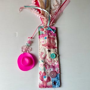 May include: A handmade bookmark with a pink cowboy hat charm. The bookmark features a patchwork design with pink, blue, and white fabrics, adorned with buttons, ribbons, and a cowboy boot illustration. Various ribbons and lace extend from the top.