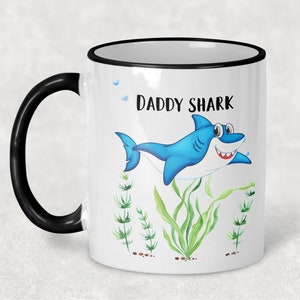 May include: White ceramic mug with a black rim and handle. The mug features a blue cartoon shark with the text "Daddy Shark" printed on the front. The shark is surrounded by green seaweed.