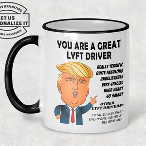 May include: White ceramic coffee mug with a black rim and handle. The mug features a cartoon illustration of a man with blonde hair and a red tie. The text on the mug reads "You are a great Lyft driver. Really terrific, quite fabulous, unbelievable, very special, huge heart, so smart. Other Lyft drivers? Total disasters! Everyone agrees! Believe me!"