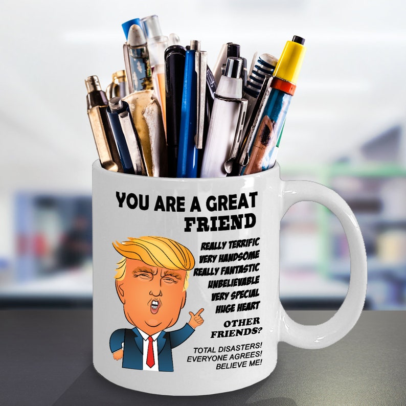 May include: White ceramic coffee mug with a cartoon image of Donald Trump. The mug has the text "You are a great friend" and a list of reasons why, including "Really terrific", "Very handsome", "Really fantastic", "Unbelievable", "Very special", "Huge heart", "Total disasters! Everyone agrees! Believe me!"