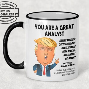May include: White ceramic coffee mug with a black rim and handle. The mug features a cartoon image of Donald Trump with the text "You are a great analyst. Really terrific, quite fabulous, unbelievable, very special, huge heart, so smart. Other analysts? Total disasters! Everyone agrees! Believe me!"