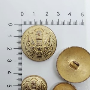 May include: Three gold-colored buttons with a design featuring a star, a building, and two flags. The buttons are on a white surface with a ruler.
