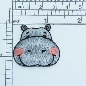 May include: A gray embroidered iron-on patch of a cartoon hippopotamus with pink cheeks. The hippopotamus is smiling with its mouth closed.