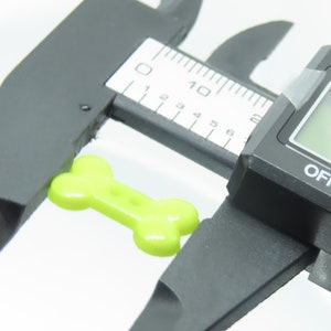 May include: A lime green, bone-shaped object is measured with a digital caliper. The caliper displays a measurement of 1.81 inches. The caliper is black with red, yellow, and blue buttons.