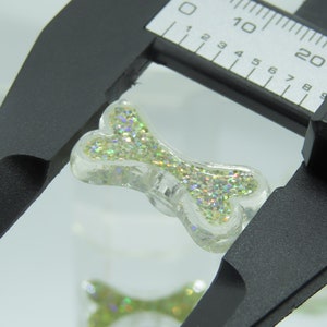 May include: A clear, glitter-filled dog bone charm is measured with a digital caliper. The bone is filled with iridescent glitter, creating a sparkling effect. The caliper shows a measurement of approximately 0.75 inches.