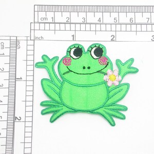 May include: Green embroidered frog applique with pink and yellow flower. The frog has big eyes and a wide smile.