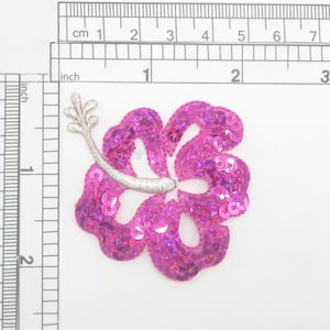 Może przedstawiać: Aplikacja z różowej kwiatu hibiskusa z cekinami, ze srebrnym łodygą i liśćmi. Kwiat ma około 5 cm szerokości.