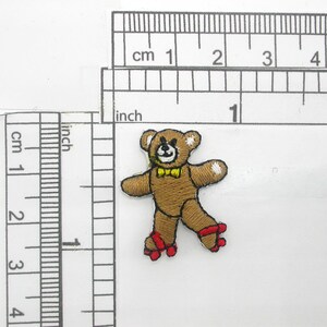 May include: Brown teddy bear wearing a yellow bow tie and red roller skates. The bear is approximately 5 cm tall.